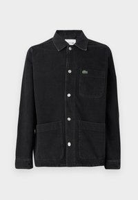 Giacca in denim nera con colletto, bottoni argentati, due tasche frontali e un piccolo logo verde Lacoste sulla tasca del petto.