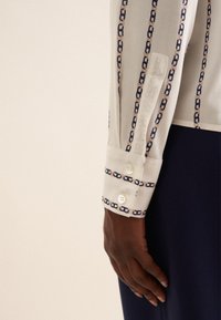 Weißes Blusenshirt mit einem gemusterten Design, das navyfarbene Kettenlinks und beigefarbene Akzente aufweist. Die Manschette hat zwei weiße Knöpfe und eine glatte Textur.