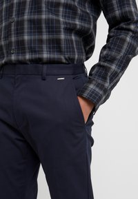 Marinblå byxor i en slät tyg, med en sidoficka och en liten metallisk logotyp som accent vid midjan. Kombinerat med en rutmönstrad skjorta i grått och blått.