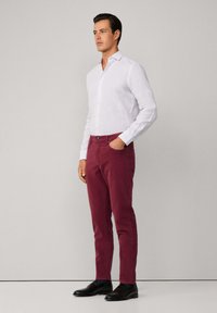 Witte linnen blouse met lange mouwen, bordeaux katoenen broek en zwarte leren schoenen; model staat zijwaarts met handen in de zakken.