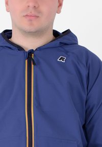 Giacca blu con zip e cappuccio, caratterizzata da una striscia verticale colorata al centro e un piccolo logo sul petto sinistro. Texture liscia.