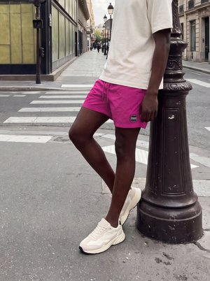 Person iført hvid skjorte, lyserøde shorts og hvide sneakers, der læner sig op ad en lamppost på en byfortov nær et fodgængerfelt.