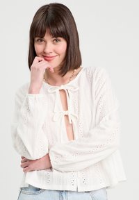 Blouse blanche en tissu texturé, avec un décolleté en V profond, des liens à l'avant et de longues manches bouffantes. Des motifs en œillets ajoutent du détail.