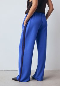 Kék sportnadrág laza szabással, keskenyedő bokarész és kontrasztos bordó csíkokkal az oldalak mentén. Puha anyag textúra.