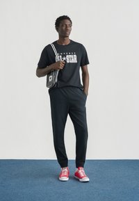 Μαύρο κοντομάνικο μπλουζάκι με εκτύπωση «CONVERSE ALL-STAR», συνδυασμένο με μαύρο παντελόνι και κόκκινα αθλητικά παπούτσια. Αξεσουάρ περιλαμβάνουν μια μικρή μαύρη τσάντα.