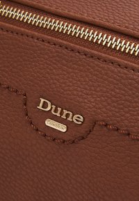 Brązowa torba z teksturowanej skóry z złotym zamkiem błyskawicznym, wytłoczonym logo „Dune” i detalami przeszyć. Kompaktowy design, odpowiednia na codzienne użytkowanie.