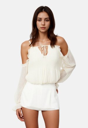 Giovane donna con capelli lunghi e scuri che indossa una camicetta bianca con scollo a barca e maniche trasparenti, abbinata a pantaloncini bianchi, in piedi davanti a uno sfondo semplice.