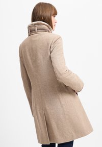 Beige uldfrakke med skræddersyet design, høj krave med pelsfoer og subtilt tekstureret mønster. Bagfra vises sømdetaljer og lommer.