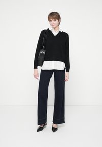 Jeune personne debout, portant un pull noir à col en V, une chemise blanche à col, un pantalon large noir, des talons noirs et portant un sac à bandoulière noir.