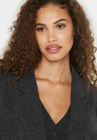 Gray blazer with a textured fabric and lapels, featuring a V-neckline shirt underneath. Loose, curly hair frames the face.