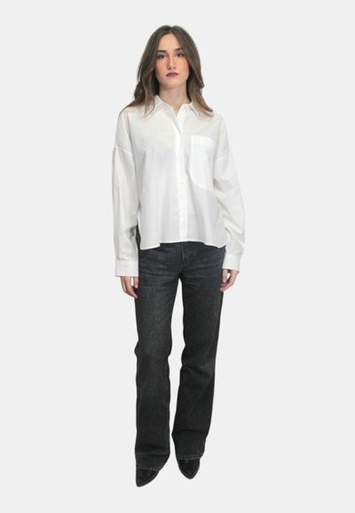 Camicia bianca a maniche lunghe con bottoni e taschino sul petto, abbinata a jeans a zampa d'elefante in denim blu scuro e décolleté neri a punta.