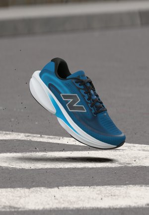 Chaussure de running New Balance bleue et noire suspendue au-dessus de l'asphalte avec des marquages routiers blancs, montrant un mouvement dynamique et des détails.