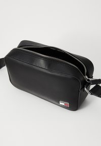 Svart lær crossbody-veske med doble glidelåslommer, slank rektangulær form og sølvfarget hardware. Inkluderer en liten logo-lapp.
