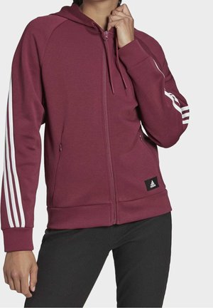 Osoba ubrana w bordową bluzę z kapturem na zamek błyskawiczny, z białymi paskami na rękawach i logo Adidas w pobliżu dołu, w połączeniu z czarnymi spodniami.