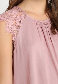 Světle růžový top s krátkými rukávy, který má složité krajkové detaily na ramenou a na výstřihu shromážděnou látku pro větší texturu.