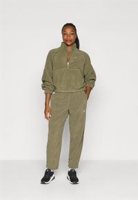 Olijfgroen fleece trainingspak met een ritskraag, een voorzak, een elastische taille en bijpassende broek. Zwarte sportschoenen maken de outfit compleet.