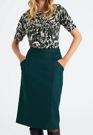 Femme portant un haut à manches courtes à motifs et une jupe mi-longue taille haute vert foncé avec les mains dans les poches.