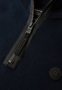 Námořnický svetr na zip s texturovanou pleteninou, černý zip s táhlem a kruhová nášivka loga na levé straně.