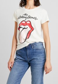 Bílý bavlněný tričko s výrazným červeným a černým logem Rolling Stones, které obsahuje grafiku rtů a jazyka. Spárováno s modrými džínami.