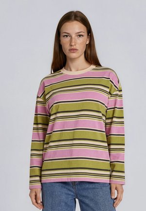 Jeune femme aux longs cheveux bruns portant un t-shirt à manches longues rayé rose et vert et un jean bleu, face à un fond uni.
