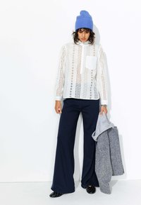 Blouse blanche à motifs avec découpes, pantalon large bleu marine, bonnet en tricot bleu, tenant un pull gris. Chaussures noires à pois blancs.