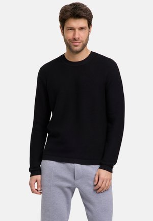 Schwarzer Strickpullover mit Rundhalsausschnitt, ausgestattet mit gerippten Bündchen und Saum. Der Stoff wirkt weich und strukturiert und wird mit hellgrauen Hosen kombiniert.