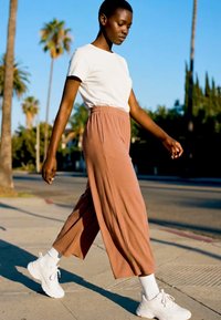 Fiatal személy sétál egy napsütéses járdán, fehér pólót, rozsdaszínű bőszárú nadrágot, fehér zoknit és fehér tornacipőt viselve.