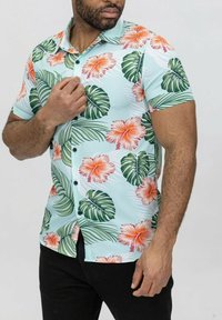 Chemise à manches courtes boutonnée de couleur bleu clair, ornée de motifs floraux et de feuilles audacieux en vert, orange et rose, avec des boutons noirs et un tissu lisse.