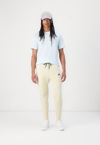 Hellblaues T-Shirt mit kurzen Ärmeln, cremefarbene Jogginghosen mit seitlichen Taschen, elastischem Bund und schwarzem Nike-Logo, kombiniert mit weißen Sneakers und einer grauen Kappe.