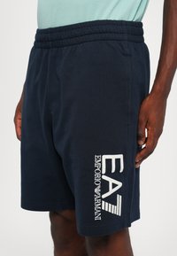Námornícke modré šortky s elastickým pásom, s veľkým bielym vyšitým logom "EA7" a textom "EMPORIO ARMANI" na ľavej strane.