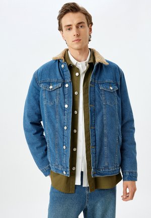 Giacca di denim con colletto in sherpa, di colore blu, con tasche frontali e chiusura con bottoni, indossata sopra una camicia verde e una camicia bianca.
