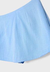 Tissu bleu clair avec une texture subtile et des coutures, probablement une partie d'un vêtement comme une jupe ou un short sur fond blanc.