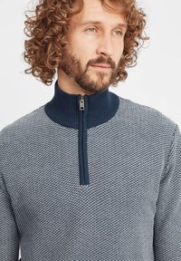 Pulover tricotat în albastru marin și alb, având un guler înalt cu fermoar, un model texturat și manșete cu nervuri. Material ușor.