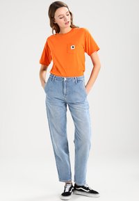 Orange bomullströja med ficka, kombinerad med blå högmidjade jeans och svarta skate-skor. Enkel, avslappnad outfit.
