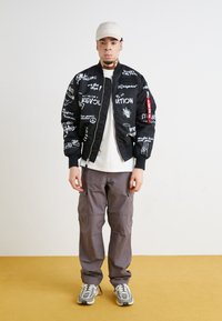 Black bomber jacket with white graphic text, zip closure, ribbed cuffs, gray cargo pants, and gray sneakers with white accents. Beige cap.
