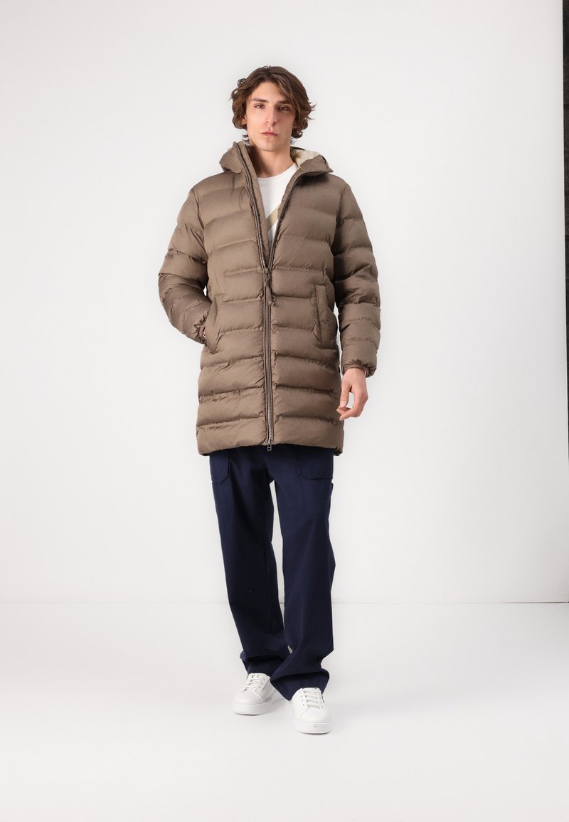 Braune Pufferjacke mit Reißverschluss vorne, hohem Kragen und gesteppten Bereichen. Kombiniert mit navyblauen Hosen und weißen Sneakern.