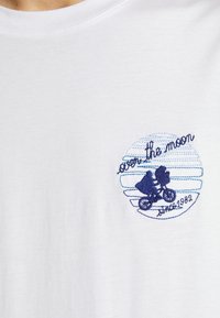 T-shirt blanc avec un motif brodé bleu d'une personne à vélo volant devant un cercle rayé avec le texte « over the moon since 1982 ».
