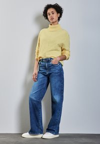 Gelber Rollkragenpullover mit strukturiertem Muster, kombiniert mit weit geschnittenen blauen Jeans und weißen Sneakers. Das Model steht vor einem schlichten Hintergrund.