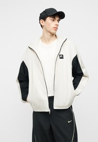 Νεαρός άνδρας με λευκό και μαύρο μπουφάν Nike Air με φερμουάρ, μαύρο παντελόνι Nike, λευκό πουκάμισο και μαύρο καπέλο, με τα χέρια στις τσέπες, κοιτάζοντας πλάγια.