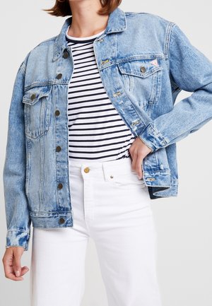 Woman wearing light blue denim jacket over black and white striped shirt with white pants, hand resting on hip.