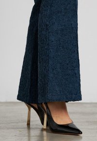Zapatos de tacón alto de charol negro con punta afilada y tacón de aguja transparente, combinados con pantalones anchos de pierna ancha de textura azul oscura.