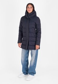 Marineblauwe gewatteerde jas met capuchon, horizontaal gestikte patroon, ritsluiting aan de voorkant en zijzakken. Gecombineerd met losvallende denim jeans en witte sneakers.