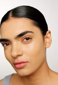 Close-up of a face with smooth, light tan skin, dark eyebrows, and full pale pink lips. Hair is slicked back, background is light gradient.