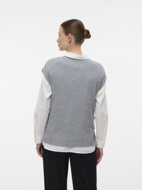 Moteris su sušukuotu plauku, dėvinti sidabrinius auskarus, baltą ilgomis rankovėmis marškinį, pilką be rankovių megztinį ir juodas kelnes, atsukusi nugarą į baltą foną.