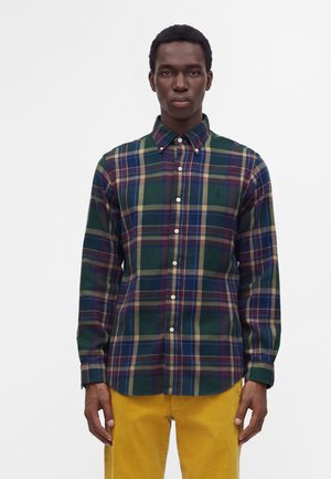 Homme portant une chemise à carreaux verte, bleue et violette boutonnée avec un pantalon jaune, debout devant un fond blanc uni.