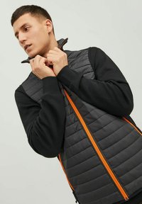Padded Weste mit schwarzem Steppmuster und orangefarbenen Reißverschlüssen. Die Ärmel sind aus schwarzem Stoff und verfügen über gerippte Bündchen. Nahaufnahme des Kragenbereichs.