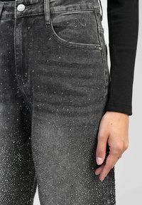 Jean en denim noir avec une texture délavée, ornés de clous en argent, comportant une poche avant et des coutures décoratives près de la taille.