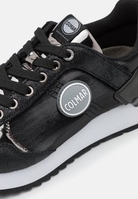 Černé tenisky s lesklým a semišovým povrchem, vybavené kulatým stříbrným logem, černými šněrovadly a bílou mezipodešví. Klasický sportovní design.