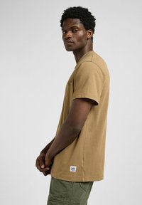T-shirt in cotone marrone con maniche corte, scollatura rotonda, taschino sul petto e una piccola etichetta del marchio sull'orlo inferiore.
