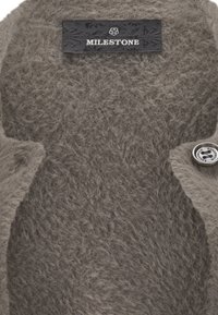 Nahaufnahme eines flauschigen, grauen Strickstoffs mit einem schwarzen Etikett, auf dem "MILESTONE" steht, und einem metallischen Knopf auf der rechten Seite.